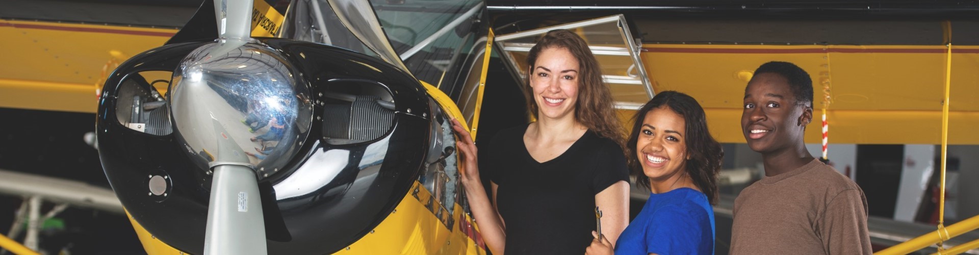 Three older youth (aged around 16) smile while standing next to a small plane